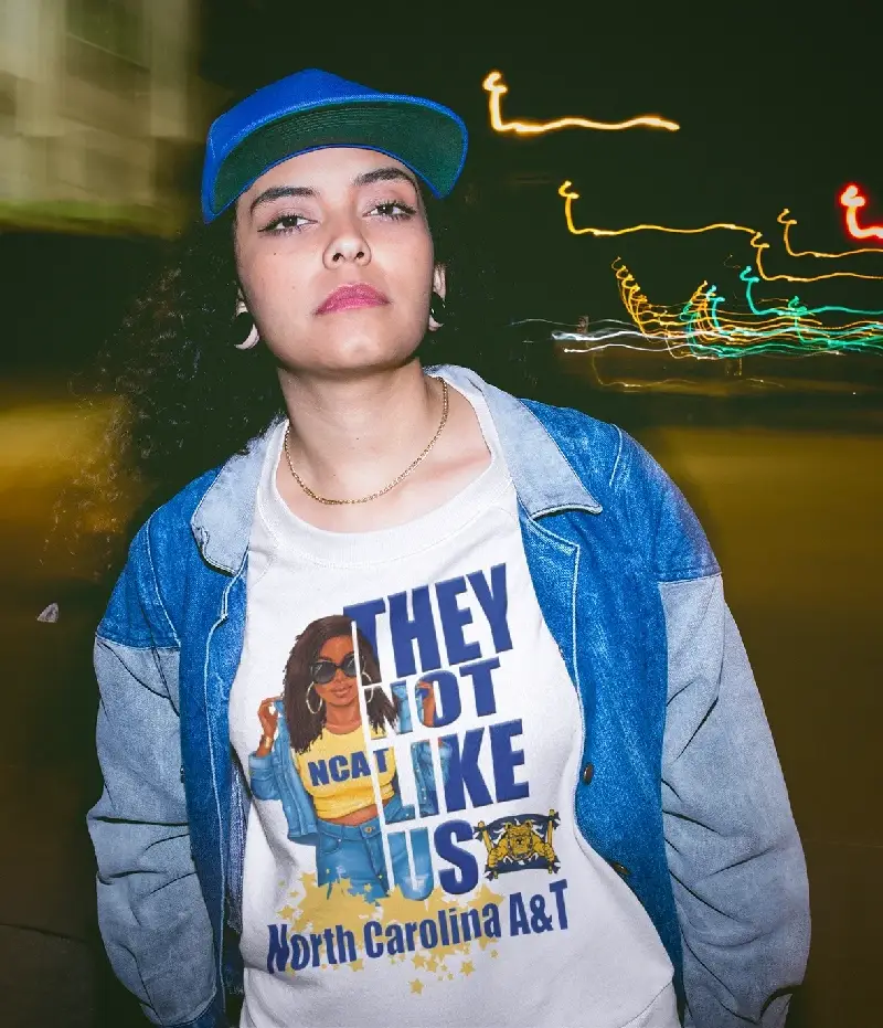 A person wearing a denim jacket and a North Carolina A&T t-shirt stands against a blurred urban backdrop at night