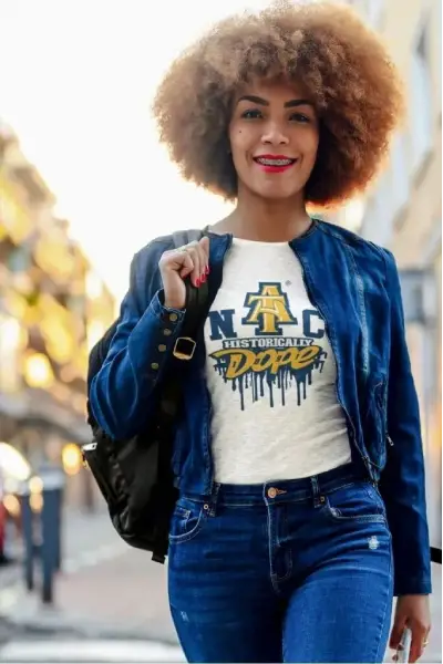 A woman with curly hair walks confidently in a blue denim jacket and graphic tee, carrying a backpack in a vibrant urban setting