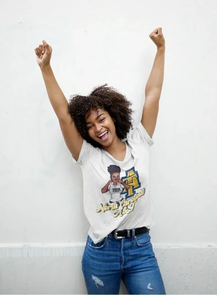 imagejpeg_0-2c753ea A person raises their arms in celebration, wearing a light grey t-shirt with "North Carolina A&T" and graphic design, against a white wall.